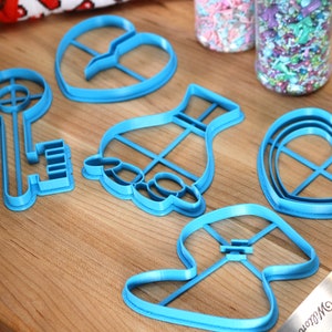 May include: A set of six blue plastic cookie cutters in various shapes, including a key, a heart, a vase, and a mushroom. The cutters are on a wooden surface.