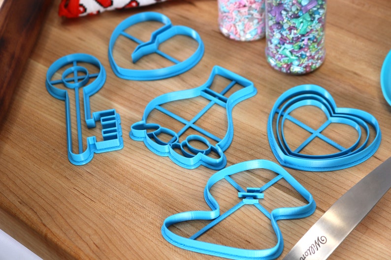 May include: A set of six blue plastic cookie cutters in various shapes, including a key, a heart, a vase, and a mushroom. The cutters are on a wooden surface.