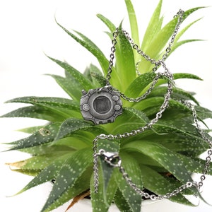 Puede incluir: Un collar de plata con un colgante con forma de personaje de videojuego. El colgante es de forma circular con un cuadrado en el centro y una superficie texturizada. El collar est&aacute; sobre una planta verde.