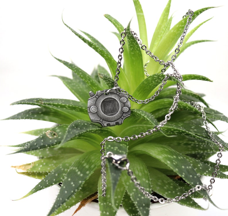 Puede incluir: Un collar de plata con un colgante con forma de personaje de videojuego. El colgante es de forma circular con un cuadrado en el centro y una superficie texturizada. El collar est&aacute; sobre una planta verde.