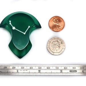 May include: A dark green, abstract-shaped object with a white line design, possibly a decorative piece. A penny and a coin are next to it for size comparison. A ruler shows measurements in inches.