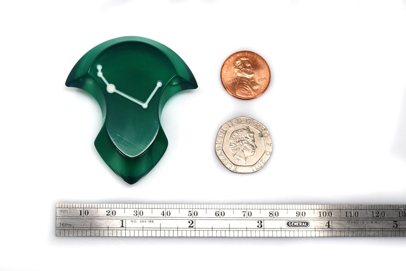 May include: A dark green, abstract-shaped object with a white line design, possibly a decorative piece. A penny and a coin are next to it for size comparison. A ruler shows measurements in inches.