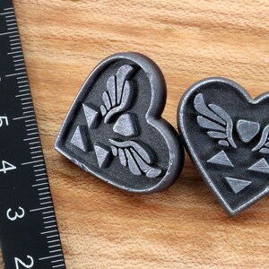Puede incluir: Dos colgantes de plata en forma de coraz&oacute;n con un dise&ntilde;o alado estilizado y un s&iacute;mbolo triangular.