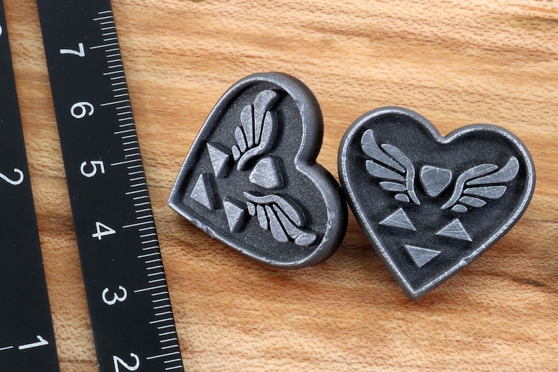 Puede incluir: Dos colgantes de plata en forma de coraz&oacute;n con un dise&ntilde;o alado estilizado y un s&iacute;mbolo triangular.