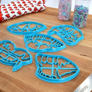 May include: A set of five blue plastic cookie cutters shaped like lips, a mountain range, and a crab. The cutters are on a wooden surface with sprinkles in jars and a blue spatula.