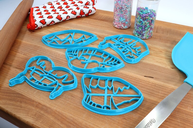 May include: A set of five blue plastic cookie cutters shaped like lips, a mountain range, and a crab. The cutters are on a wooden surface with sprinkles in jars and a blue spatula.