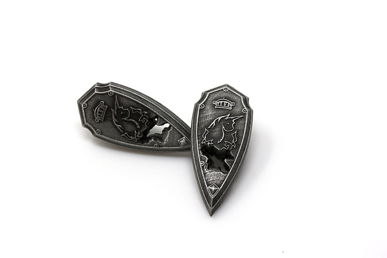 May include: Two silver-toned metal shield-shaped pins with a unicorn design. The unicorn is facing left on one pin and right on the other. The pins are on a white background.