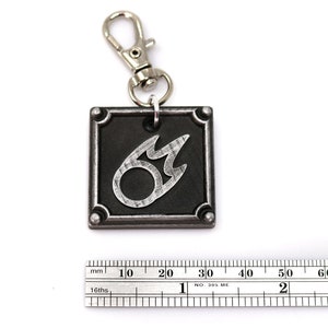 May include: A silver and black metal keychain featuring the Final Fantasy VII Fire Materia symbol. The keychain is attached to a silver split ring.