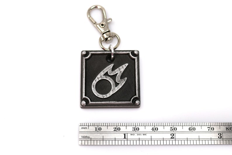 May include: A silver and black metal keychain featuring the Final Fantasy VII Fire Materia symbol. The keychain is attached to a silver split ring.