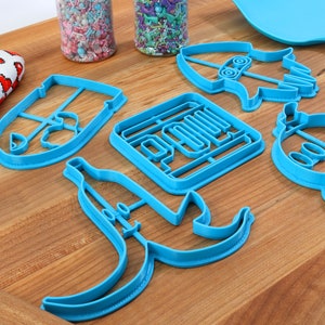 May include: A set of six blue plastic cookie cutters shaped like a banana, a bottle, a square with the word "POUM" inside, a rocket, a robot, and a shield. The cutters are on a wooden surface.