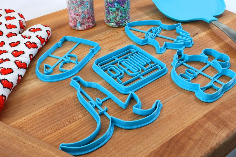 May include: A set of six blue plastic cookie cutters shaped like a banana, a bottle, a square with the word "POUM" inside, a rocket, a robot, and a shield. The cutters are on a wooden surface.