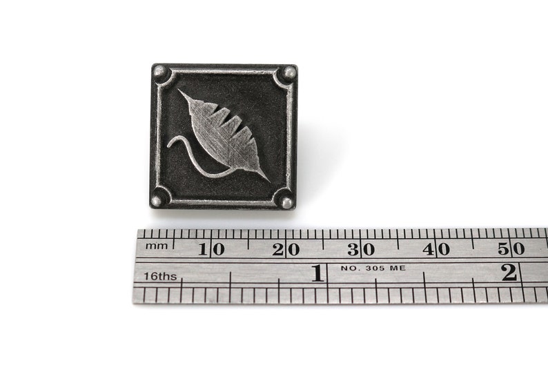 May include: A silver metal square with a stylized leaf design. The square is approximately 1.5 inches wide.
