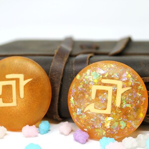 May include: Two round, orange resin bookmarks with a white kanji character on each. The bookmarks are on a brown leather journal with a leather strap.