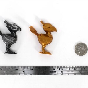 May include: Two 3D printed figurines of a mythical creature, one in black and one in gold. The figurines are about 5 cm tall and are sitting on a white surface. A ruler is in the background.