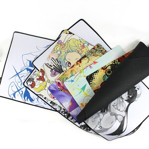 May include: A stack of colorful mousepads with various anime and graphic designs. The mousepads are on a white surface. One mousepad is rolled up and has a black surface.