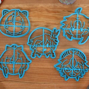 Pu&ograve; includere: Cinque tagliabiscotti in plastica blu a forma di volti di cartoni animati. I tagliabiscotti sono su una superficie di legno.