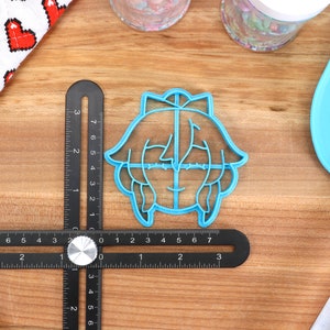Pu&ograve; includere: Un tagliapasta in plastica blu a forma di personaggio dei cartoni animati con un fiocco sulla testa. Il tagliapasta &egrave; su una superficie di legno con un righello e una spatola blu.