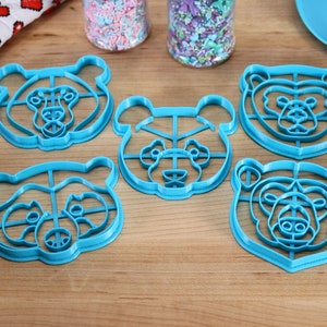 May include: Five blue plastic bear-shaped cookie cutters on a wooden surface.