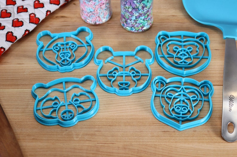 May include: Five blue plastic bear-shaped cookie cutters on a wooden surface.
