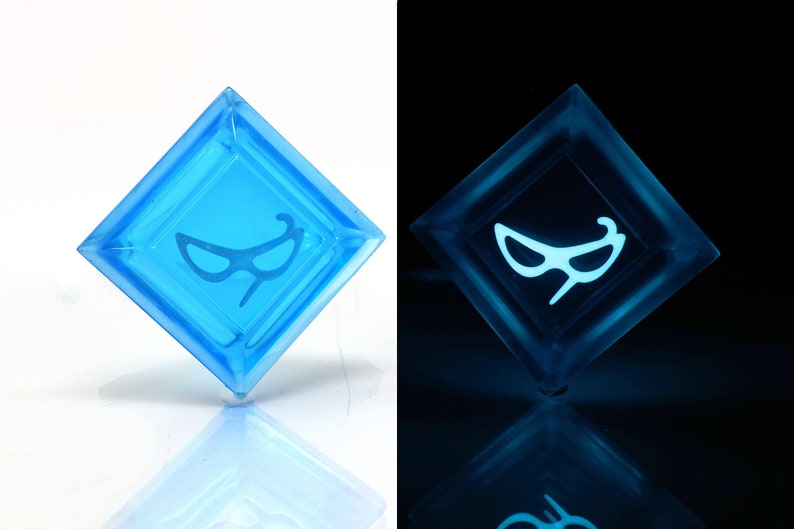 May include: A blue, diamond-shaped, glow-in-the-dark keycap with a white, stylized image of a pair of sunglasses.