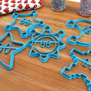 May include: A set of six blue plastic cookie cutters shaped like a star, a sun, a crescent moon, a sheriff's badge, a pixelated spaceship, and a pixelated character.