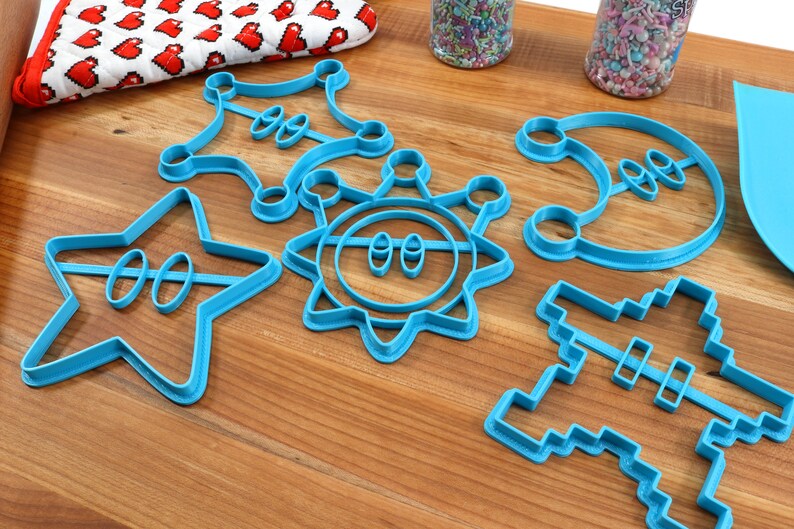 May include: A set of six blue plastic cookie cutters shaped like a star, a sun, a crescent moon, a sheriff's badge, a pixelated spaceship, and a pixelated character.
