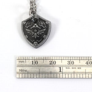 May include: A silver necklace with a pendant shaped like a shield. The shield has a black and silver design with a stylized bird and a triangle. The pendant is about 2 cm wide.