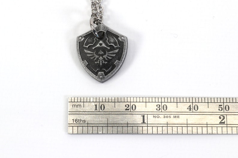 May include: A silver necklace with a pendant shaped like a shield. The shield has a black and silver design with a stylized bird and a triangle. The pendant is about 2 cm wide.
