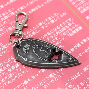 May include: A silver metal keychain featuring a shield-shaped charm with a black and white unicorn design. The charm has a distressed, aged look and a star-shaped hole in the center. The keychain has a silver metal clasp.