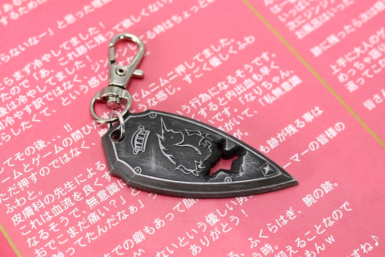 May include: A silver metal keychain featuring a shield-shaped charm with a black and white unicorn design. The charm has a distressed, aged look and a star-shaped hole in the center. The keychain has a silver metal clasp.