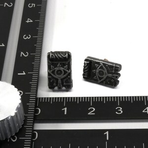 May include: Two black metal earrings with a silver design of an eye with triangles around it. The earrings are on a white surface with a black ruler.