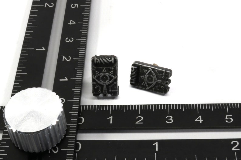 May include: Two black metal earrings with a silver design of an eye with triangles around it. The earrings are on a white surface with a black ruler.