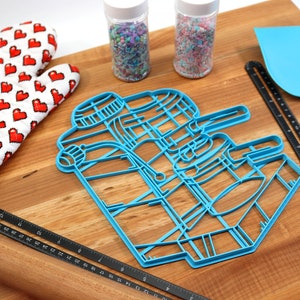 May include: A blue plastic cookie cutter shaped like a robot on a wooden surface with a rolling pin, a red oven mitt, and two jars of sprinkles.