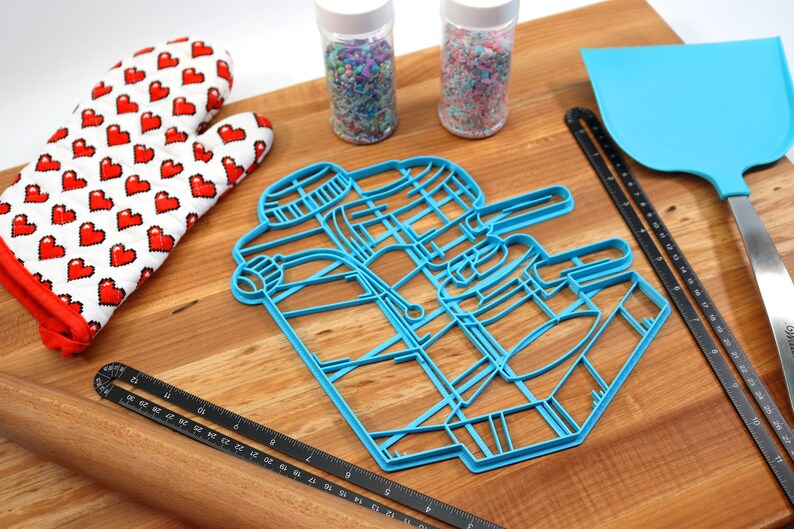 May include: A blue plastic cookie cutter shaped like a robot on a wooden surface with a rolling pin, a red oven mitt, and two jars of sprinkles.