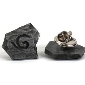 Puede incluir: Un pin de piedra gris con un dise&ntilde;o en espiral, con un cierre de metal.