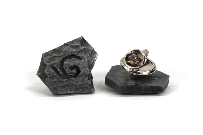 Puede incluir: Un pin de piedra gris con un dise&ntilde;o en espiral, con un cierre de metal.