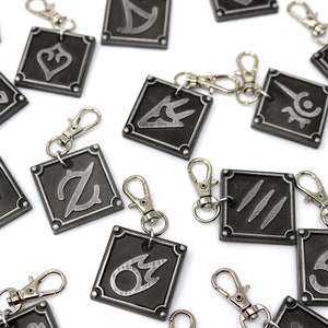 May include: A set of 15 silver and black metal keychains with different symbols. Each keychain features a square-shaped charm with a unique symbol in a silver color on a black background.