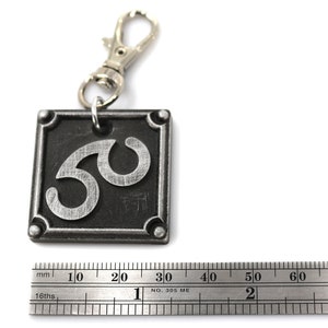 May include: A silver metal keychain with a black square charm featuring the number 50 in a stylized font. The charm has a silver metal ring attached to a silver metal lobster clasp.