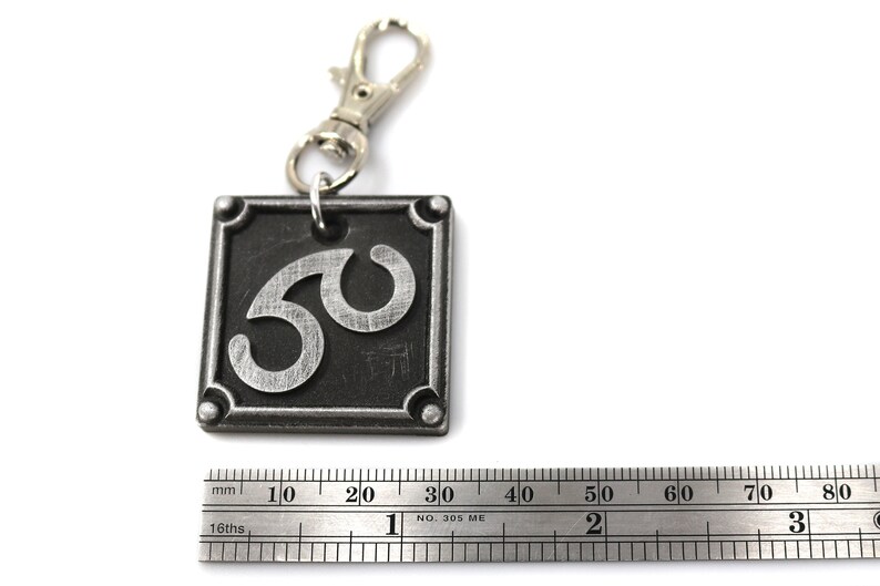 May include: A silver metal keychain with a black square charm featuring the number 50 in a stylized font. The charm has a silver metal ring attached to a silver metal lobster clasp.