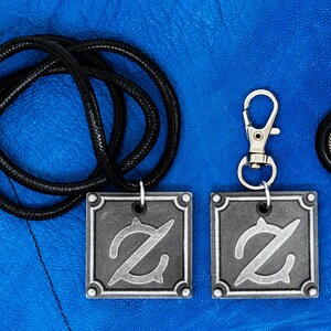 Pu&ograve; includere: Due ciondoli in metallo color argento con una lettera Z stilizzata su una collana con cordoncino nero e un portachiavi in metallo color argento con una lettera Z stilizzata. I ciondoli e il portachiavi sono su uno sfondo blu.