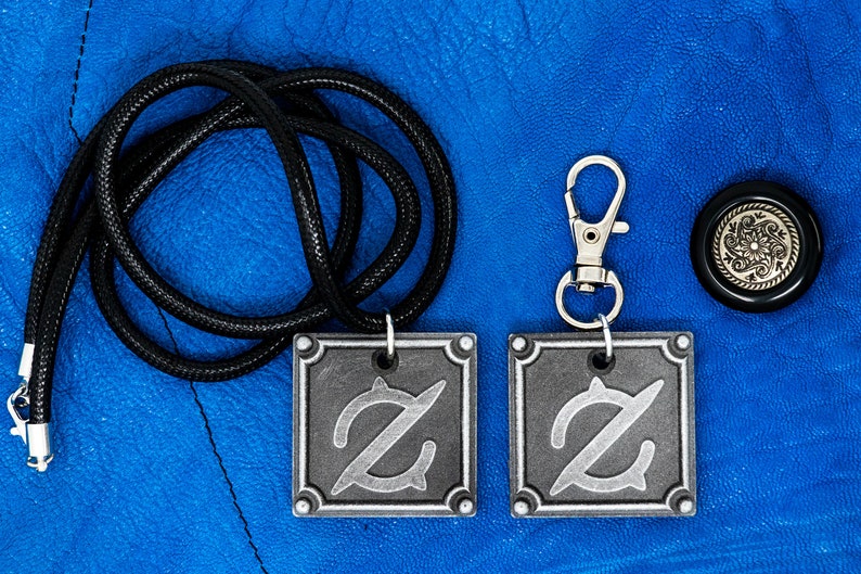 Pu&ograve; includere: Due ciondoli in metallo color argento con una lettera Z stilizzata su una collana con cordoncino nero e un portachiavi in metallo color argento con una lettera Z stilizzata. I ciondoli e il portachiavi sono su uno sfondo blu.
