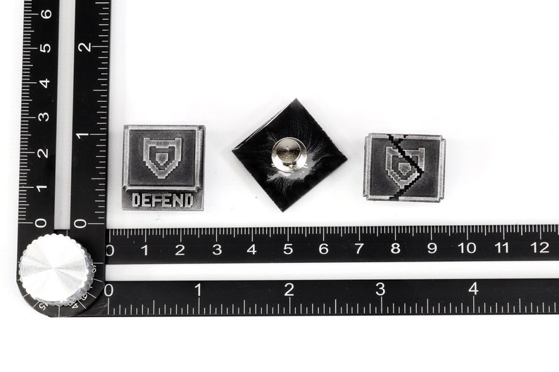 May include: Three silver-colored metal pins with a pixelated shield design. The first pin has the word "DEFEND" printed below the shield. The second pin is a blank back. The third pin has a crack through the shield.