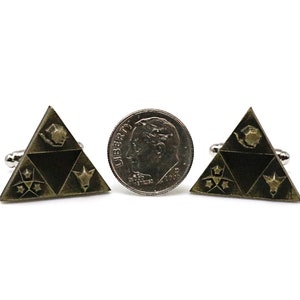 May include: A pair of gold-toned cufflinks shaped like a triangle with a black background. The triangle features four symbols, including a star, a crescent moon, a bird, and a stylized animal head. The cufflinks are shown next to a US dime for scale.