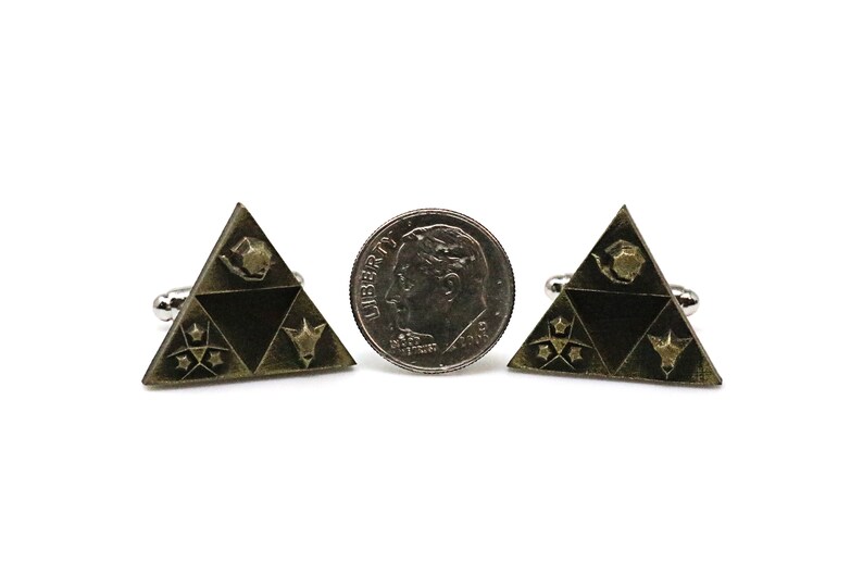 May include: A pair of gold-toned cufflinks shaped like a triangle with a black background. The triangle features four symbols, including a star, a crescent moon, a bird, and a stylized animal head. The cufflinks are shown next to a US dime for scale.