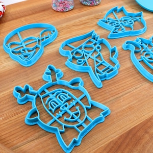 Peut inclure: Six emporte-pi&egrave;ces en plastique bleu clair repr&eacute;sentant des personnages de dessins anim&eacute;s, dont un lion, un cheval et un robot. Un gant de cuisine rouge et blanc &agrave; motifs de c&oelig;urs est visible en arri&egrave;re-plan.