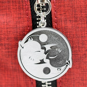 May include: A silver yin yang charm with a black and white design of two animals facing each other. The charm is attached to a silver zipper pull.