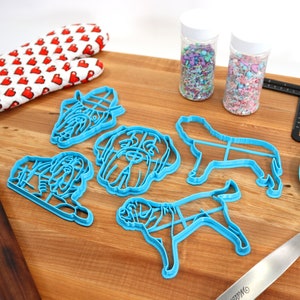 Peut inclure: Un ensemble d'emporte-pi&egrave;ces en forme de chien en plastique bleu, un rouleau &agrave; p&acirc;tisserie en bois, un gant de cuisine rouge &agrave; c&oelig;urs blancs, deux pots de vermicelles color&eacute;s, une spatule bleue et un outil de mesure noir sur une surface en bois.