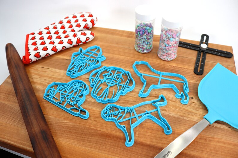 Peut inclure: Un ensemble d'emporte-pi&egrave;ces en forme de chien en plastique bleu, un rouleau &agrave; p&acirc;tisserie en bois, un gant de cuisine rouge &agrave; c&oelig;urs blancs, deux pots de vermicelles color&eacute;s, une spatule bleue et un outil de mesure noir sur une surface en bois.