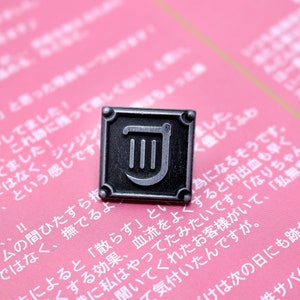 Op de afbeelding: Een zilveren metalen pin met een zwarte vierkante achtergrond en een zilveren liersymbool. De pin is op een roze oppervlak met Japanse tekst.