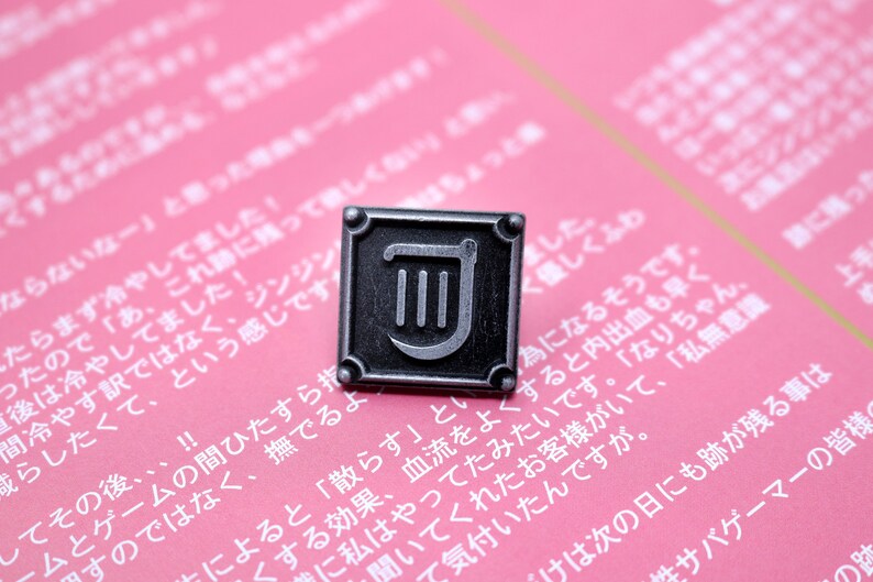 Op de afbeelding: Een zilveren metalen pin met een zwarte vierkante achtergrond en een zilveren liersymbool. De pin is op een roze oppervlak met Japanse tekst.